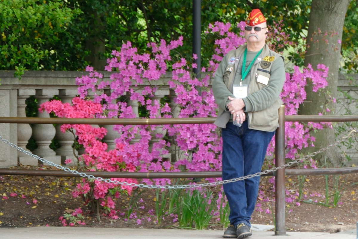 Twin Tiers' chaplain leaning against railing with blooming flowers behind.