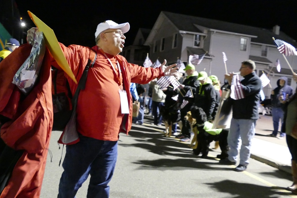 Man standing with open arms out with crowd waving american flags in the background at a mission homecoming.